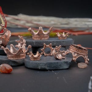 May include: A set of seven rose gold crown-shaped charms, some with open centers, arranged on gray stone surfaces.