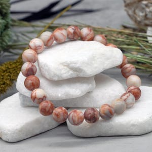 May include: A beaded bracelet featuring round, reddish-brown and cream-colored stones. The bracelet is displayed on white, textured, stone-like surfaces, with greenery in the background. The beads appear to be approximately 0.5 inches in diameter.