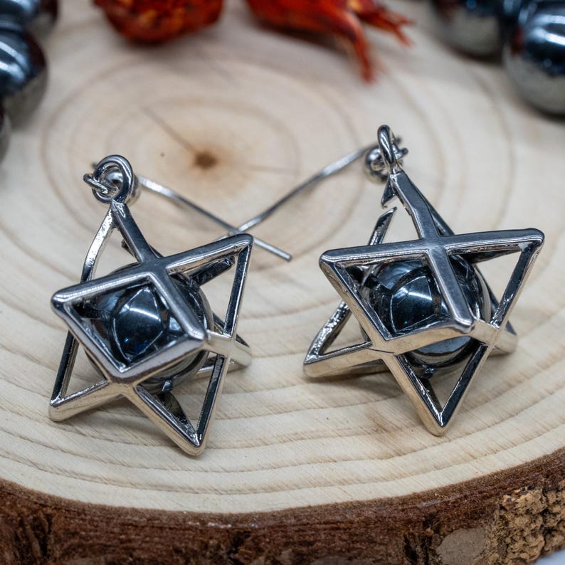 May include: A pair of silver earrings. Each earring has a geometric design with a star shape holding a dark, spherical bead. The earrings are shown on a wooden surface, with other decorative elements in the background.