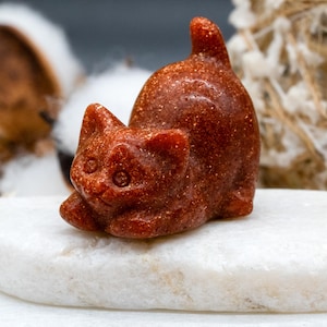 May include: A small, orange-brown, carved cat figurine made of sunstone. The cat is sitting on a white surface.