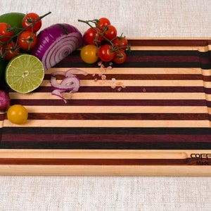 May include: A striped wooden cutting board with a red onion, lime, and tomatoes on top. The board has a dark wood border and the text "COFFMAN Made in USA" is printed on the bottom right corner.