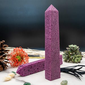 Op de afbeelding: Twee paarse, obeliskvormige decoratieve items met een textuuroppervlak. Eén staat rechtop, de andere ligt horizontaal. De achtergrond bevat een dennenappel, een rood-oranje bloem en een vetplant. De items staan op een wit oppervlak.
