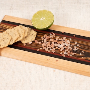 May include: A wooden serving board with a black, red, and brown striped design. The board is topped with a lime wedge, pink Himalayan salt, and crackers.
