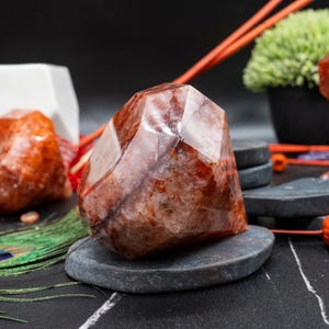 May include: A faceted, reddish-brown crystal geode sits on a gray stone. The crystal has a geometric shape with multiple facets, displaying variations in color and texture. The background includes decorative elements and a black surface.
