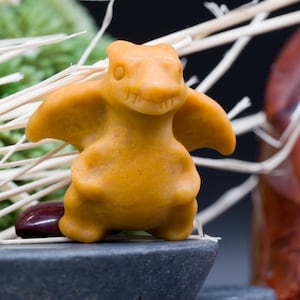 Può includere: Una piccola statuina giallo-arancio di una creatura alata, forse un drago o un dinosauro, con le ali spiegate. La statuina è posta su una pietra grigio scuro, con uno sfondo sfocato di vegetazione ed elementi decorativi.