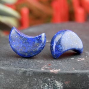 Puede incluir: Un par de pendientes en forma de media luna. Los pendientes están hechos de una piedra azul oscuro con motas doradas. Los pendientes están montados sobre postes de plata. Se exhiben sobre una superficie gris oscura.