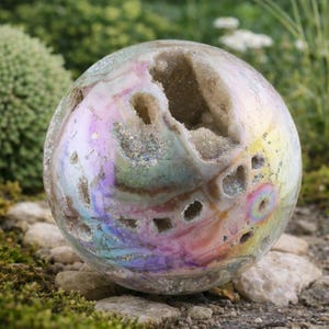 May include: An iridescent geode sphere with a rainbow sheen and several openings revealing a crystalline interior. The crystal ball sits on small rocks and moss, with greenery in the background. A decorative item.