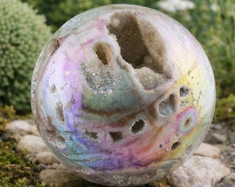 May include: A spherical, iridescent geode with a rainbow sheen. The crystal ball has several openings revealing a crystalline interior. The sphere sits on a bed of small rocks and moss, with greenery in the background.