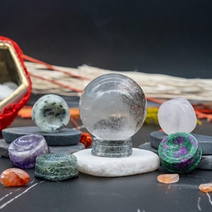 Puede incluir: Una colección de varias esferas y orbes de piedras preciosas pulidas, incluyendo una esfera de cristal de cuarzo transparente sobre una base de piedra verde, una esfera verde y morada, una esfera rosa y una esfera verde y negra. Las esferas están dispuestas sobre una superficie gris oscura.