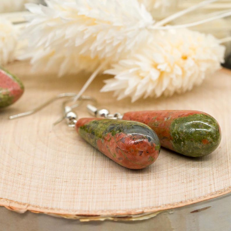 May include: A pair of drop earrings featuring polished, teardrop-shaped unakite stones with mottled green and pink patterns. The stones are suspended from silver-toned metal hooks, showcasing a smooth, glossy finish and natural mineral variations.