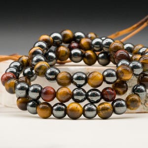 May include: Three beaded bracelets. The bracelets feature a combination of polished, round beads in shades of brown, gold, and silver. The beads are strung together, creating a layered effect. The bracelets are displayed on a light-colored surface.