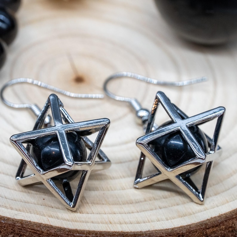 May include: A pair of silver-tone earrings featuring a geometric star design. Each earring has a black, spherical bead encased within a star-shaped frame. The earrings hang from silver hooks.