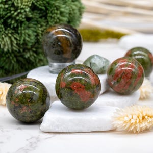 May include: Five green and red gemstone spheres on a white surface. The spheres are polished and have a speckled appearance.