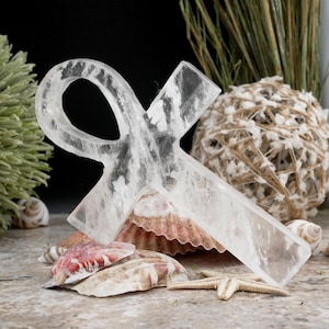 May include: A clear quartz crystal ankh, a symbol of life, rests on seashells and a starfish. The ankh is translucent with visible internal patterns. The background includes decorative elements like a woven ball and dried plants.