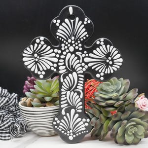 May include: Black ceramic cross with white floral designs. The cross has a hole at the top for hanging. The cross is decorated with white floral patterns and dots. The cross is displayed in front of a variety of succulents.