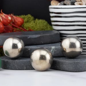 May include: Three polished, silver-coloured metal spheres sit on dark grey, rectangular stones. The spheres reflect the surrounding environment. The background includes a black and white striped pot, red floral accents, and green moss.