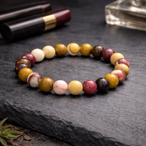 May include: A beaded bracelet featuring a variety of earth-toned beads in shades of cream, yellow, brown, and burgundy. The bracelet is displayed on a dark gray surface, with a blurred background including lipstick and a perfume bottle.