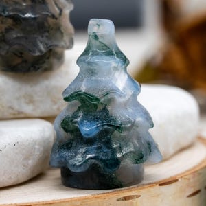 May include: A small, carved gemstone tree figurine. The tree is a light blue color with green and dark blue inclusions, resembling a miniature evergreen. The figurine is displayed on a wooden surface with white stones in the background.