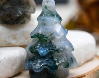 May include: A small, carved gemstone tree figurine. The tree is a light blue color with green and dark blue inclusions, resembling a miniature evergreen. The figurine is displayed on a wooden surface with white stones in the background.