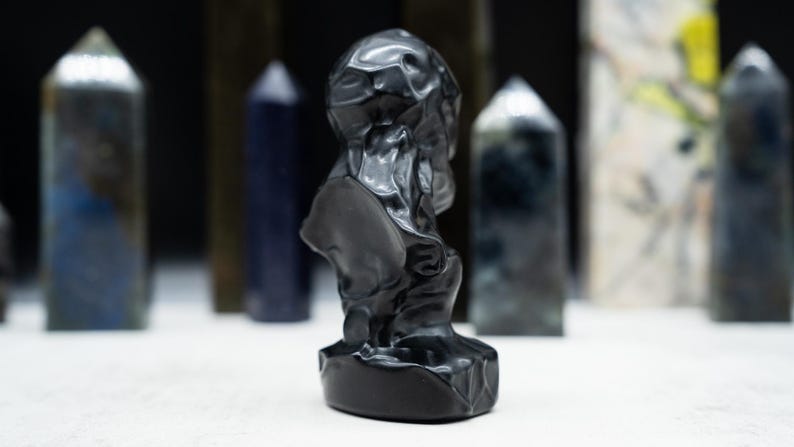 May include: A black sculpture of a bust with a textured surface, set against a blurred background of various dark-colored, pointed crystal formations. The sculpture is the focal point, with the crystals adding depth.