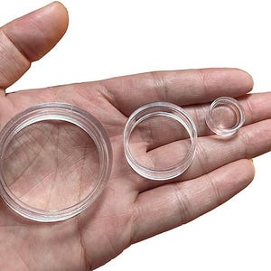May include: Three clear, circular plastic coin holders of varying sizes are displayed. The largest holder is approximately 2 inches in diameter, the medium one is about 1.25 inches, and the smallest is around 0.75 inches. The holders are transparent, allowing a clear view of any coin placed inside.
