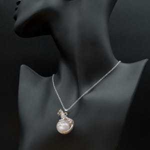 Pode incluir: Um colar prateado com um pingente que retrata um leopardo abraçando uma grande pérola redonda e perolada. O colar é exibido em um manequim preto. A corrente é delicada.