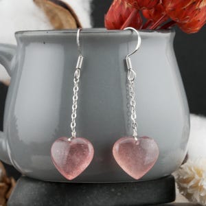 Puede incluir: Pendientes colgantes con gemas rosas translúcidas en forma de corazón. Los pendientes tienen ganchos y eslabones plateados. Las gemas tienen un aspecto moteado. Los pendientes se muestran sobre un fondo gris.