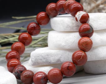 May include: A red jasper beaded bracelet. The bracelet features large, round beads in varying shades of red with unique patterns. The beads are strung together, forming a complete circle. The bracelet is displayed on white, textured stones.