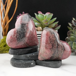 May include: Two heart-shaped stones with pink and black marbling sit atop a stack of gray heart-shaped stones. The background includes green succulents and a black and white striped pot, with a black backdrop.