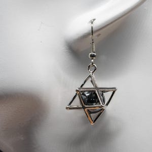May include: A single silver earring with a geometric star design. The earring has a wire hook and a small, dark, spherical bead encased within the star's framework. The earring is shown against a white background.