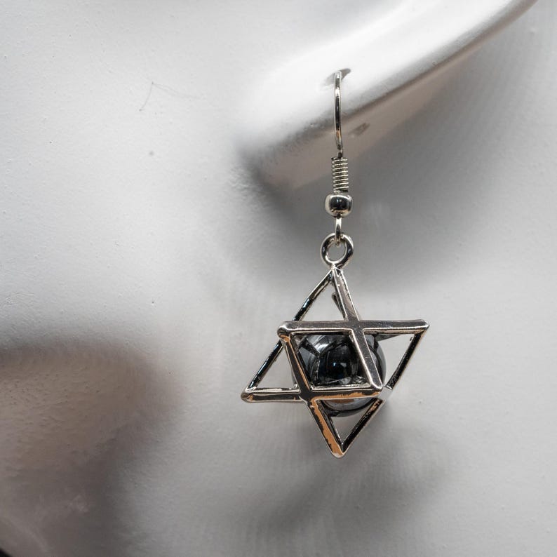 May include: A single silver earring with a geometric star design. The earring has a wire hook and a small, dark, spherical bead encased within the star's framework. The earring is shown against a white background.