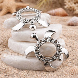 May include: Two silver-colored turtle-shaped ornaments with clear crystal accents. The ornaments are displayed on white stones, with a sandy background and seashells. The turtle's shell is a circle of crystals.