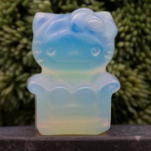 Puede incluir: Una figura translúcida de gato con un lazo, que presenta un degradado de colores de azul a amarillo. La figura con forma de gato tiene rasgos faciales visibles y está colocada sobre un fondo verde borroso.