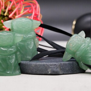May include: Three green crystal figurines of a character with large ears and a robe are displayed. One figurine is lying on its side, while the other two are upright. A black cord and a dark gray stone are also present.