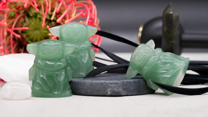 May include: Three green crystal figurines of a character with large ears and a robe are displayed. One figurine is lying on its side, while the other two are upright. A black cord and a dark gray stone are also present.
