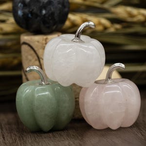 Puede incluir: Tres calabazas de cristal talladas, una blanca, una rosa y una verde. Las calabazas están dispuestas sobre una superficie de madera.
