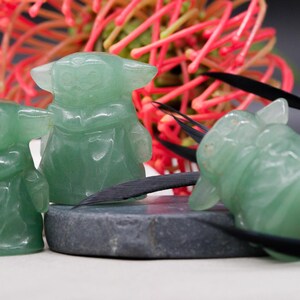 May include: Three green jadeite figurines of a character with large ears and a robe. The figurines are on a gray stone and white surface, with a red and green floral arrangement in the background. Black decorative elements are also present.