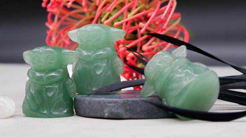 May include: Three green jadeite figurines of a character with large ears and a robe. The figurines are on a gray stone and white surface, with a red and green floral arrangement in the background. Black decorative elements are also present.