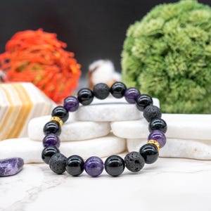 May include: A beaded bracelet featuring a combination of black, purple, and textured lava rock beads. The bracelet includes gold-colored spacers. The beads are strung together, creating a circular design. The bracelet is displayed on a white surface.