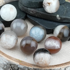 Può includere: Una collezione di sfere di pietre preziose lucide in vari colori, tra cui grigio, marrone, bianco e nero. Le sfere sono esposte su una superficie di legno, con alcune appoggiate su pietre scure e piatte. Le sfere variano in dimensioni e presentano motivi unici.