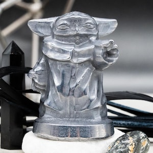 May include: A silver-colored figurine of a character with large ears and a robe, standing on a white stone. The figurine is the focal point, with black decorative elements and a black bottle in the background.