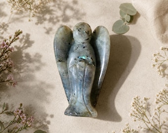 May include: A polished labradorite gemstone carved into the shape of an angel with smooth, rounded wings and a central body. The stone displays a greyish-blue base color with natural dark inclusions and subtle iridescent flashes of light.
