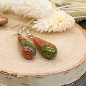 May include: A pair of drop earrings featuring polished, teardrop-shaped unakite stones with mottled green and reddish-pink patterns. The stones are suspended from silver-toned metal hooks, showcasing a smooth, glossy finish and natural color variations.