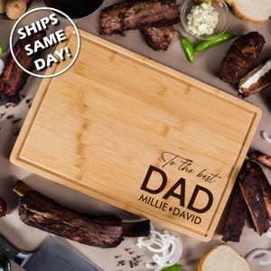 May include: A rectangular bamboo cutting board with a juice groove. The text "To the best DAD" is engraved in black, along with the names "Millie + David". The board is surrounded by cooked ribs, onions, and other food items.