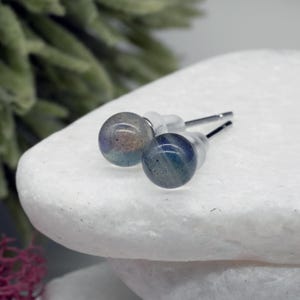 May include: A pair of stud earrings featuring round, iridescent labradorite stones. The stones display a mix of blue, green, and purple hues. The earrings have silver posts and clear backs, set against a white background.