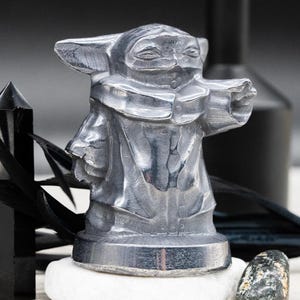 May include: A silver-colored figurine of a character from a popular science fiction franchise. The figure is standing on a white stone, with a black crystal and feathers in the background. The figurine is approximately 4 inches tall.