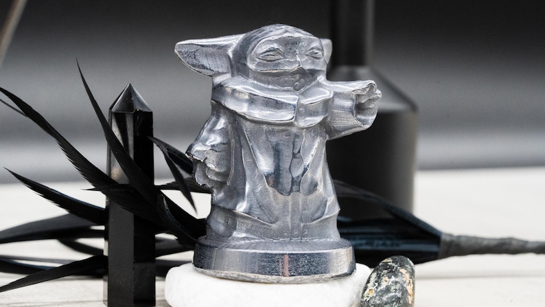 May include: A silver-colored figurine of a character from a popular science fiction franchise. The figure is standing on a white stone, with a black crystal and feathers in the background. The figurine is approximately 4 inches tall.