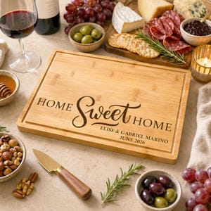 May include: A rectangular bamboo cutting board featuring a juice groove and laser-engraved text that reads HOME Sweet HOME, ELISE & GABRIEL MARINO, JUNE 2026. The board has a light wood grain finish and is positioned centrally on a flat surface.