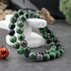 May include: A beaded bracelet featuring round, polished stones in shades of forest green, black, and occasional deep magenta. The beads are strung together in a circular formation, displaying a natural mottled texture and varied color distribution.