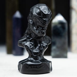 May include: A black bust sculpture of a character with a detailed face and chest. The sculpture is set against a blurred background of various crystal towers. The bust has a textured surface and is made of a dark, possibly obsidian, material.